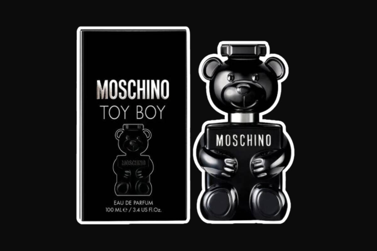O perfume Toy Boy, com seu frasco em formato de ursinho de pelúcia metalizado, com a caixa à direita, em um fundo preto
